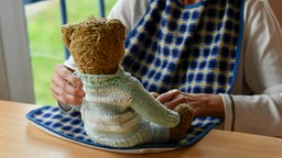 Eine Frau im höheren Alter betrachtet einen Teddybär, den sie auf einem Tisch vor sich hält.