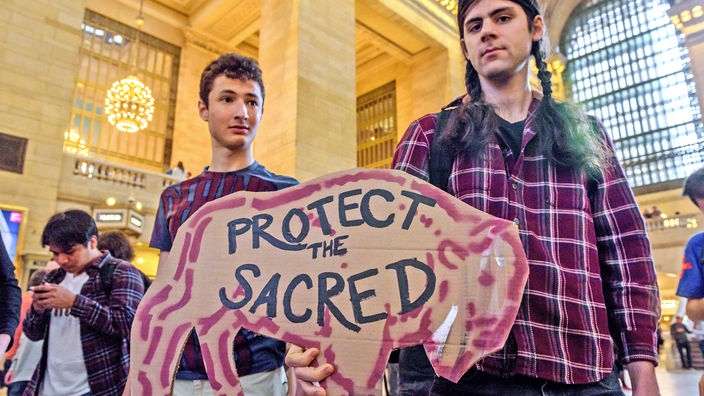 Aktivisten protestieren in New York City gegen J.P.Morgan Chase, einen der größten Investoren fossiler Brennstoffe in den USA. Dabei halten ein Schild in Form eines Bison und dem Text "protect the sacred" in die Kamera.