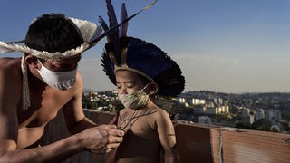Seit dem Dammbruch einer Eisenerzmiene 2019 ist die Lebensumgebung der Pataxo unbewohnbar. Teile der Volksgruppe leben seitdem in Belo Horizonte, wo sie Covid-19 bedroht. Das Bild zeigt Vater und Sohn mit Schutzmasken und traditionellem Kopfschmuck.