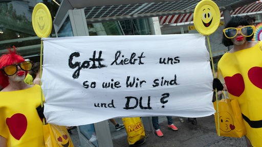 Kostümierte halten auf dem Berliner CSD ein Transparent mit der Botschaft: "Gott liebt uns so wie wir sind. Und du?"