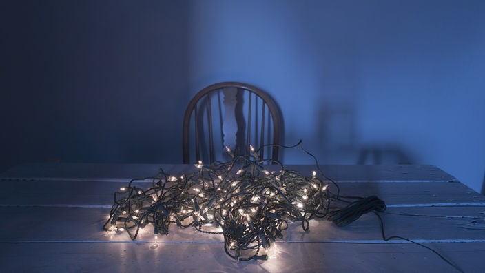 Eine Weihnachtslichterkette liegt gekräuselt als Bündel auf einem Tisch, hinter dem ein verlassener Stuhl im faden Licht steht.