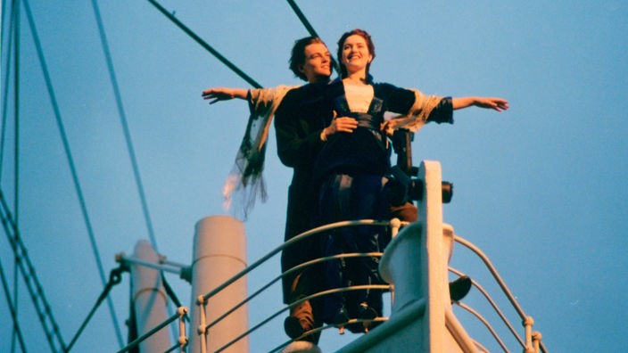 Das Bild zeigt Leonardo DiCaprio und Kate Winslet in einer Szene der Titanic Verfilmng von 1997, in der kate Winslet erwartungsvoll mit ausgebreiteten Armen an der Reling der Titanic steht, Leonardo diCaprio sie haltend dahinter.