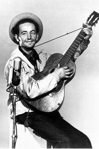 Ein Schwazweiß-Foto des Musikers Woody Guthrie, der mit einer Akustikgitarre vor einem Mikrofon steht