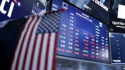Ein Bildschirm zeigt Amerikanische Aktienkurse, im Vordergrund ist eine Amerikanische Flagge zu sehen. Aufnahme vom 09.03.2020