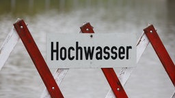 Ein Schild mit der Aufschrift "Hochwasser" ist an einer rot-weißen Straßensperre befestigt. dahinter ist unscharf eine Wasseroberfläche zu sehen