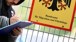 Ein Mann füllt ein Formular aus neben einem Zaun mit einem Schild des Bundesamt für Migration und Flüchtlinge