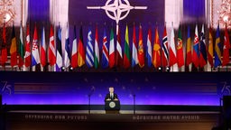 NATO Secretary General am Podium vor den Mitgliedsflaggen im prunkvollen Andrew Mellon Auditorium