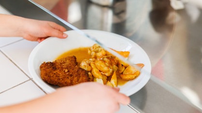 Ein Kind in einer Schulmensa nimmt einen Teller mit paniertem Fleisch und frittierten Kartoffelecken entgegen