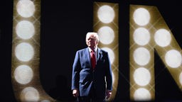 Donald Trump steht im Schlaglicht vor einer schwarzen Wand auf der Scheinwerfer seinen Namen buchstabieren