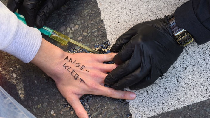 Die Hand eines Umweltaktivisten ist auf der Fahrbahn fest geklebt und ein Polizist versucht den Kleber zu lösen. 