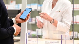 Ein Mann mit seinem Handy in der Hand, auf dem die App "Das E-Rezept" geöffnet ist, wird in einer Apotheke von einer Apothekerin bedient. 