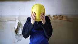 Ein Mensch in Arbeitshose hält sich einen gelben Schutzhelm vor das Gesicht.