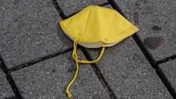 Symbolbild: Eine gelbe FFP2-Maske mit defektem Gummiband liegt auf dem Boden
