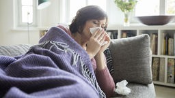 Frau liegt auf einem Sofa unter einer Decke und putzt sich die Nase. (Symbolbild) 