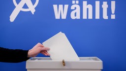 Hand wirft Umschlag in Wahlurne vor blauem Hintergrund mit der Aufschrift "Wählt!"