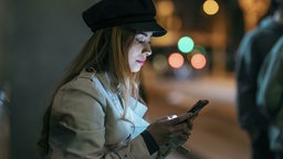 Eine junge Frau blickt nachts auf der Straße auf ihr Handy. Symbolbild