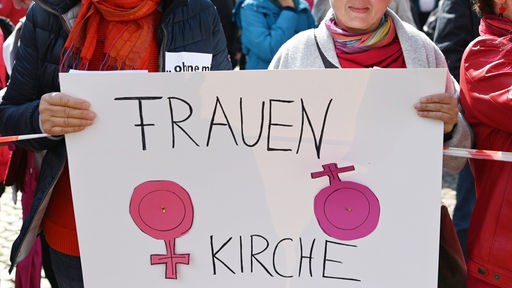 Frauen halten ein Plakat mit der Aufschrift "Frauen Kirche" und dem Symbol für Weiblichkeit, bei dem das Kreuz zusätzlich markiert ist.