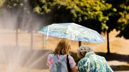 Symbolbild: Zwei Menschen, einer davon grauhaarig, gehen mit einem Regenschirm als Sonnenschutz an einem Brunnen vorbei (18.07.2022)