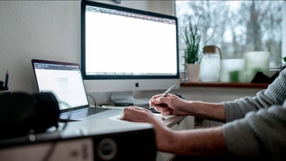 Symbolbild: Ein Mensch an einem Schreibtisch mit Laptop und Zweitbildschirm (Bildschirmansicht gerastert).