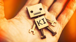 Symbol: Eine Roboterspielfigur mit Aufschrift KI liegt auf einer menschlichen Hand.