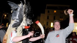 Ein rot-weiß gekleideter Mann mit riesiger Maske aus Fell und Vogelflügeln hält ein Kuhhorn in der Hand, während ein Mann mit Borkumer Logo auf dem Pullover eine Glocke läutet