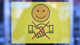 Schild an einer Glastür mit einem Schild, dass ein lächelndes Gesicht zeigt und zwei durchgestrichen Hände, die zum Gruß gereicht werden. Symbolbild
