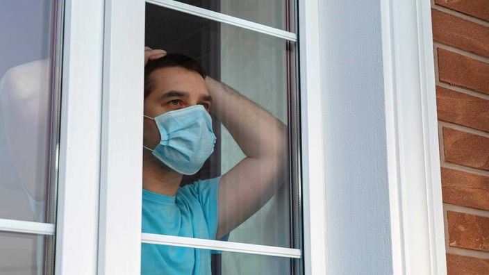 Symbolbild: Ein junger Mensch mit Corona-Schutzmaske schaut aus einem Fenster