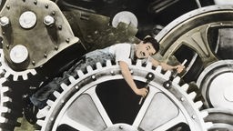 Charlie Chaplin zwischen Zahnrädern in einer Fabrik (Filmstill aus "Modern Times")