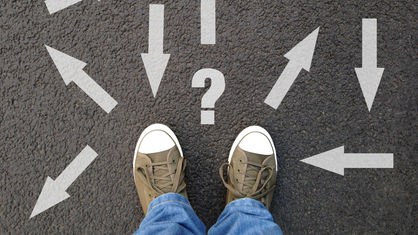 Symbolbild: Eine Person steht auf einem Boden, auf dem Pfeile in unterschiedliche Richtung sowie vor ihren Füßen ein Fragezeichen aufgedruckt ist.