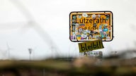 Unter dem mit  Protestfolien beklebten Ortsschild "Lützerath - Stadt Erkelenz - Kreis Heinsberg" hängt ein Schild "bleibt".