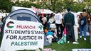 Studierende stehen zwischen Zelten. An einem davon klebt ein Banner mit der Aufschrift "Munich students stand for justice in Palestine"