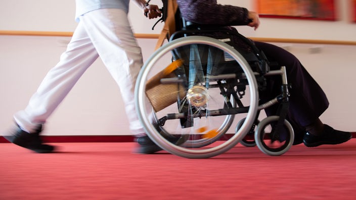 Symbolbild: Eine Pflegekraft schiebt einen Menschen im Rollstuhl