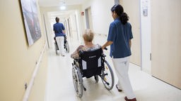 Zwei Pflegerinnen schieben Patient:innen im Rollstuhl auf einem Flur in einem Krankenhaus