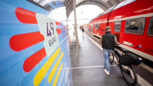 Ein Mann geht am Hauptbahnhof mit seinem Fahrrad zwischen der Werbung für das Deutschlandticket und einer Regionalbahn vorbei