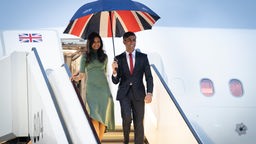 Rishi Sunak hält einen Schirm mit der britischen Falgge während er neben seiner Ehefrau aus einem Flugzeug steigt.