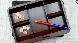 Symbolbild: Ein rot-blauer Stift liegt über einer mit wenigen Münzen gefüllten Kasse.