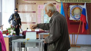 Ein Mensch wirft seinen Stimmzettel in eine Wahlurne in einem mit russischer Flagge geschmückten, von einem Soldaten bewachten Wahlraum in Luhansk (27.09.2022)