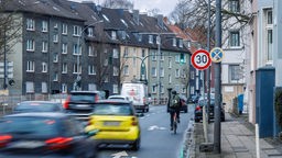 Symbolbild: Eine Straße mit Mehrfamilienhäusern am Rand, Autoverkehr, Radfahrer:innen und Tempo-30-Schild.