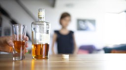 Ein gefülltes Glas und eine Flasche mit Alkohol stehen auf einem Tisch. Im Hintergrund steht ein Kind. Symbolbild 