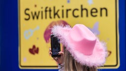 Ein Mensch mit rosa Cowboyhut vor einem Schild "Swiftkirchen".
