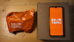 Auf einem Paket liegt ein Smartphone das die App von Temu zeigt, daneben eine orangene Versandtasche von Temu