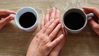 Symbolbild: Aufsicht auf zwei Hände, die sich beim Kaffeetrinken berühren
