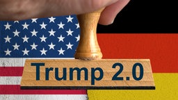 Eine Hand hält einen Stempel mit der Aufschrift "Trump 2.0". Im Hintergrund die US-Flagge und Deutsche Flagge in der Mitte zerissen. Fotomontage/Symbolbild