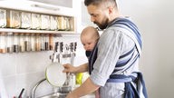 Vater mit Säugling im Tragetuch beim Geschirr spülen. Symbolbild
