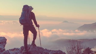 Ein Mensch mit Rucksack schaut von einem Gipfel in eine Berglandschaft