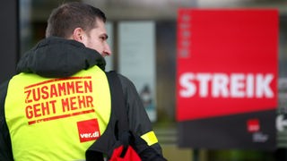 Mitarbeiter mit Warnweste steht vor einem Streikplakat