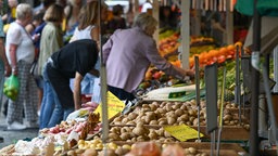 Menschen stehen an einem Stand mit Obst und Gemüse auf einem Wochenmarkt.