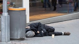 Symbolbild: Win Mensch schläft neben einem Mülleimer und seinem Pappbecher für Spenden auf der Straße