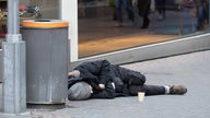 Symbolbild: Ein Mensch schläft neben einem Mülleimer und seinem Pappbecher für Spenden auf der Straße