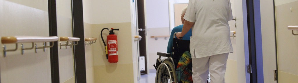 Eine Altenpflegerin schiebt einen Senior im Rollstuhl über einen Flur